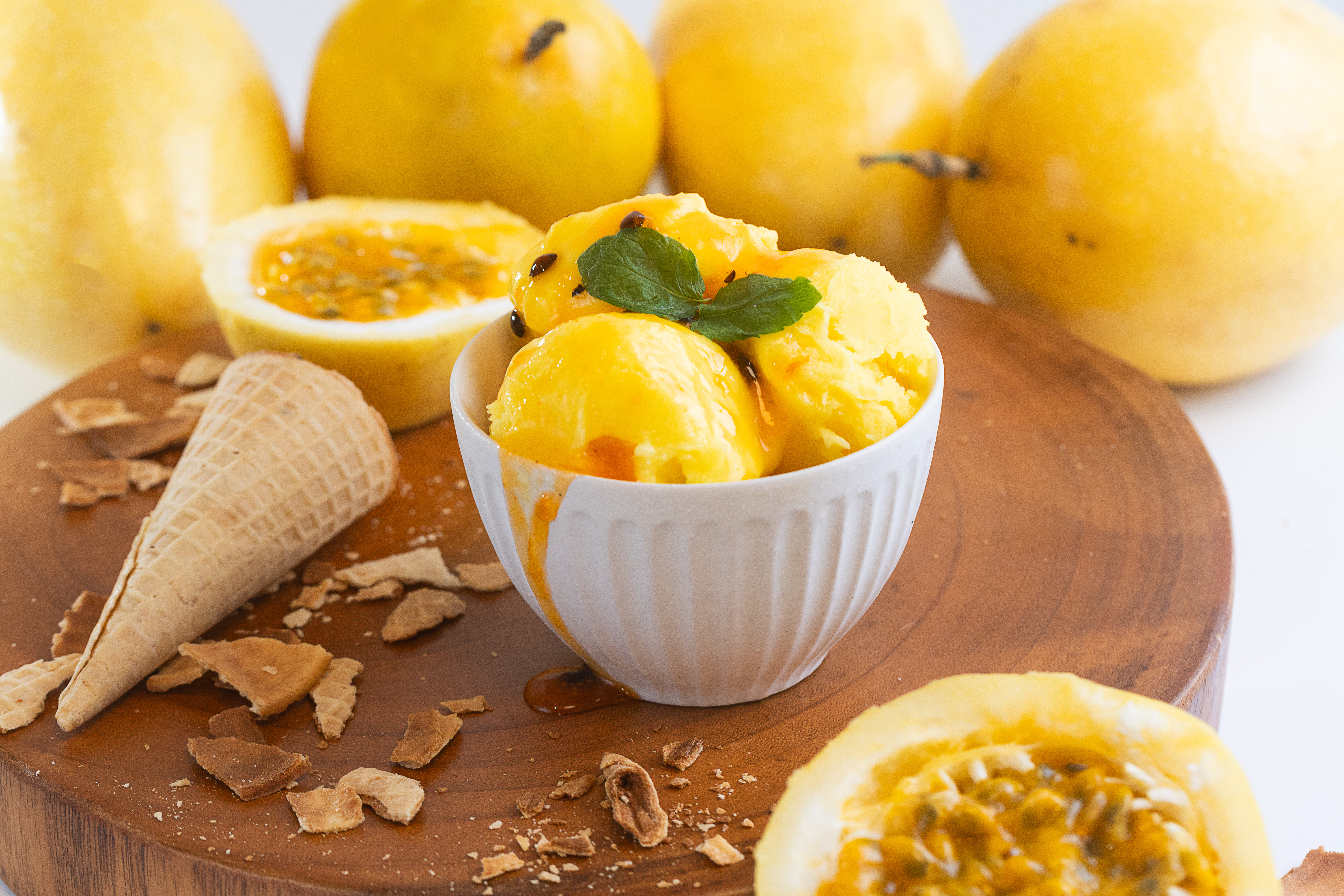 Tigela branca com bolas de sorvete de maracujá decoradas com folhas de hortelã, calda de maracujá e sementes, ao lado de casquinha quebrada e frutas de maracujá inteiras e abertas sobre base de madeira.