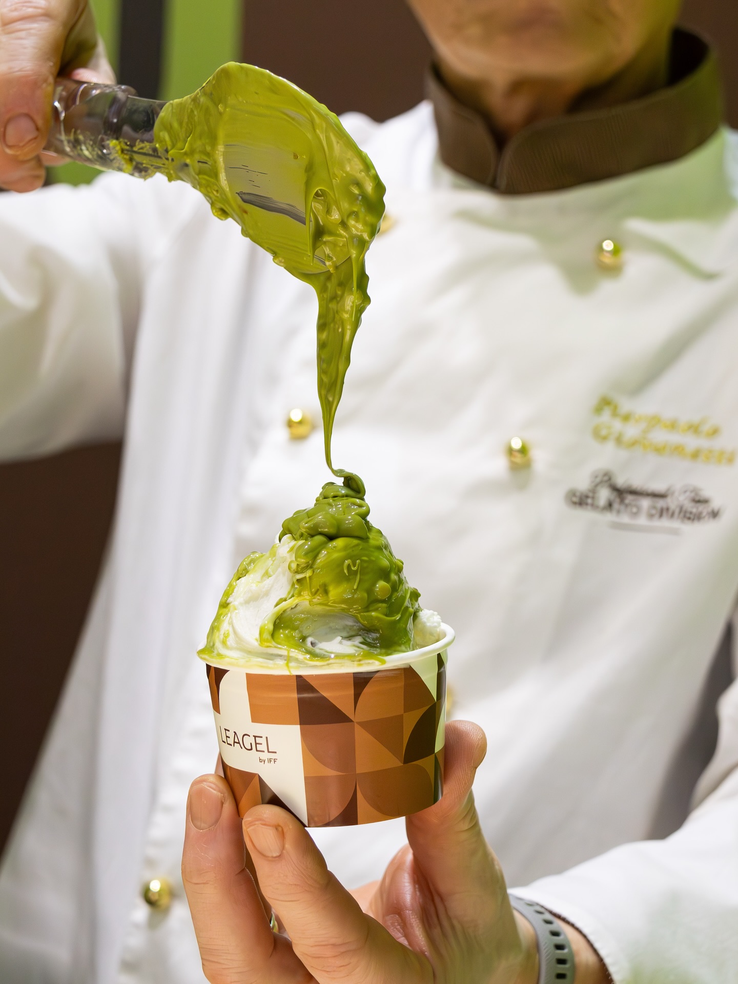  Foto de um gelatiere aplicando uma cobertura de pistache com inclusões sobre um potinho de gelato.?