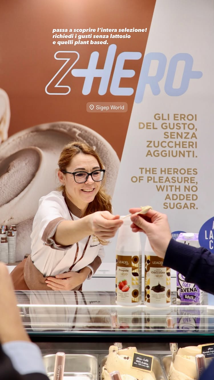 Foto de uma vitrine de gelatos sem adição de açúcar, com uma atendente oferecendo uma pazinha de degustação a um cliente.